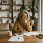 Une photographie montre une femme d'âge mûr aux longs cheveux gris ondulés et portant des lunettes, assise à une table en bois et se concentrant sur des documents.